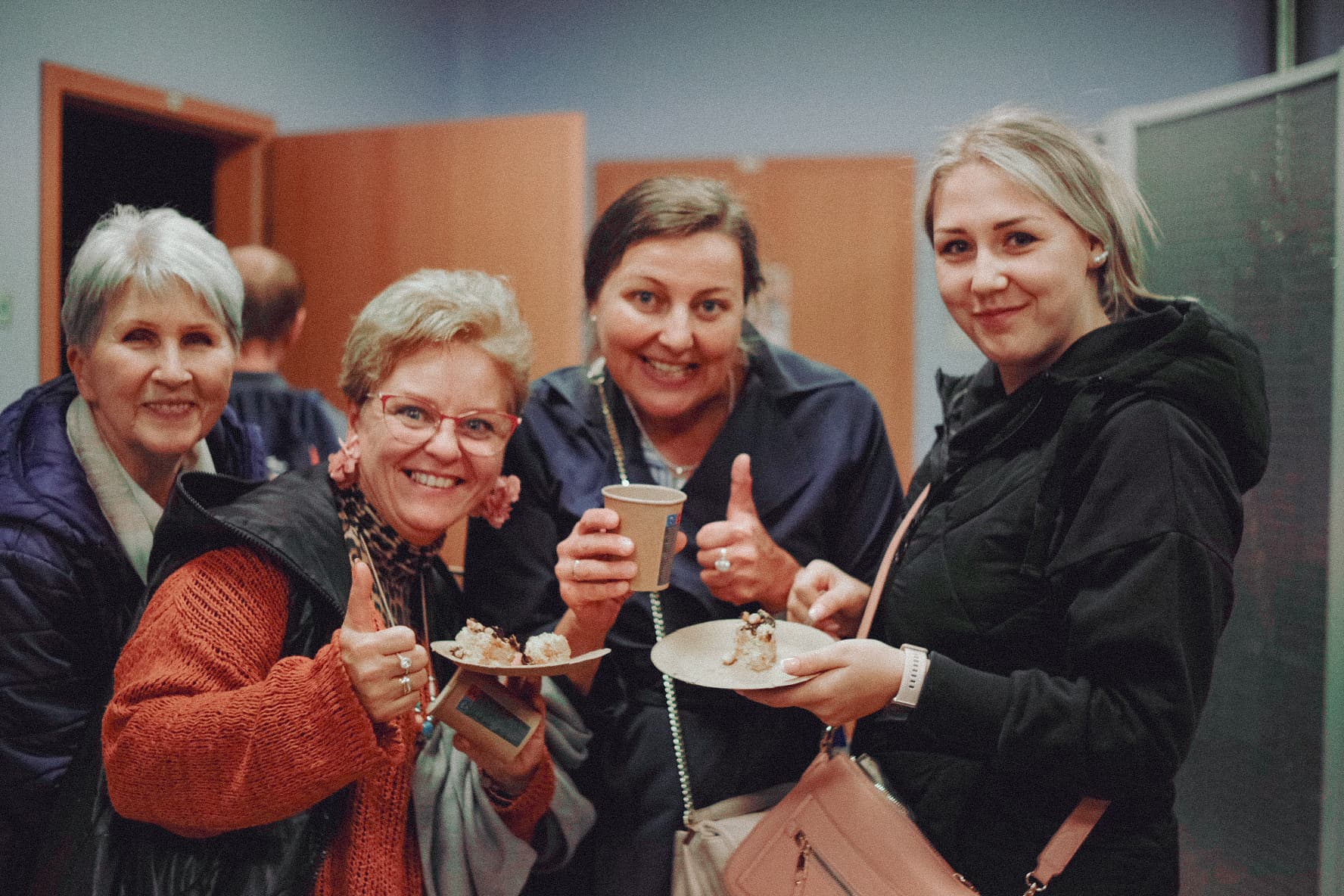 Obraz pokazuje cztery starsze kobiety siedzące razem i uśmiechające się, trzymające kubki i talerze z ciastem. Są ubrane w codzienne ubrania, a tło obejmuje wnętrze sali.