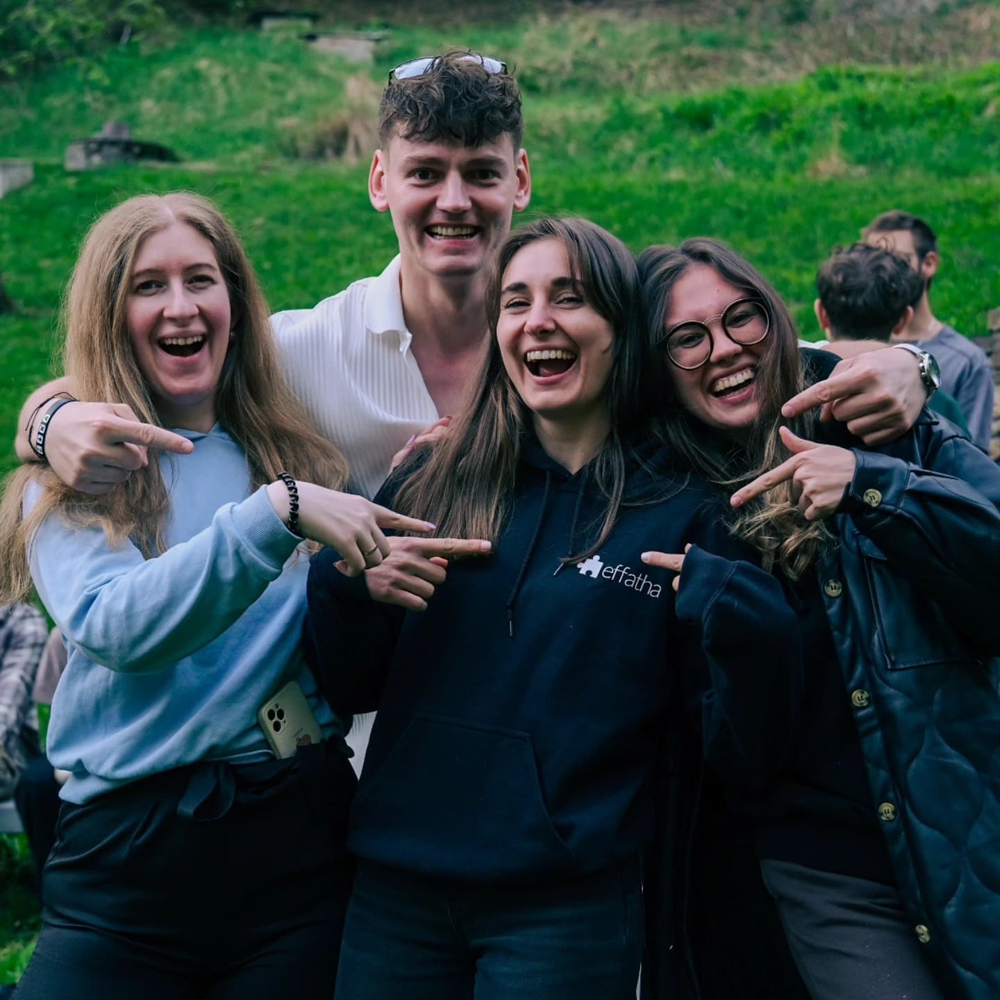 Obraz przedstawia czterech młodych ludzi stojących na trawie, uśmiechających się i wskazujących na siebie nawzajem. Są ubrani w codzienne stroje, w tym bluzy i koszulki, a tło stanowi zielone wzgórze.