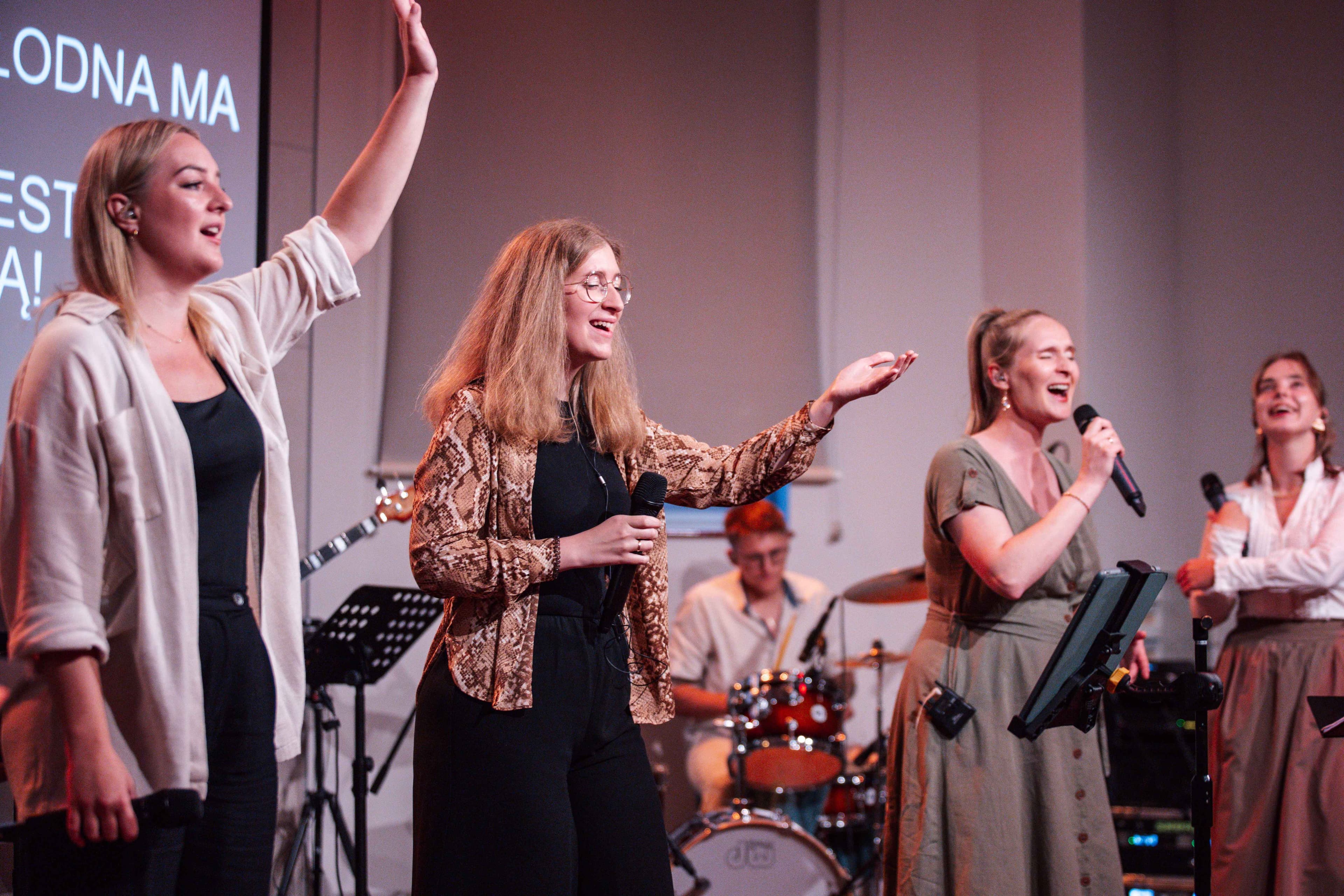 Obraz przedstawia grupę muzyków na scenie z kolorowym oświetleniem. Dwie wokalistki stoją przy mikrofonach, ubrane w jasne stroje, podczas gdy inni grają na instrumentach, takich jak skrzypce, gitara i keyboard. W tle widoczny jest ekran projekcyjny, a scena jest wyposażona w statywy na nuty i krzesła.