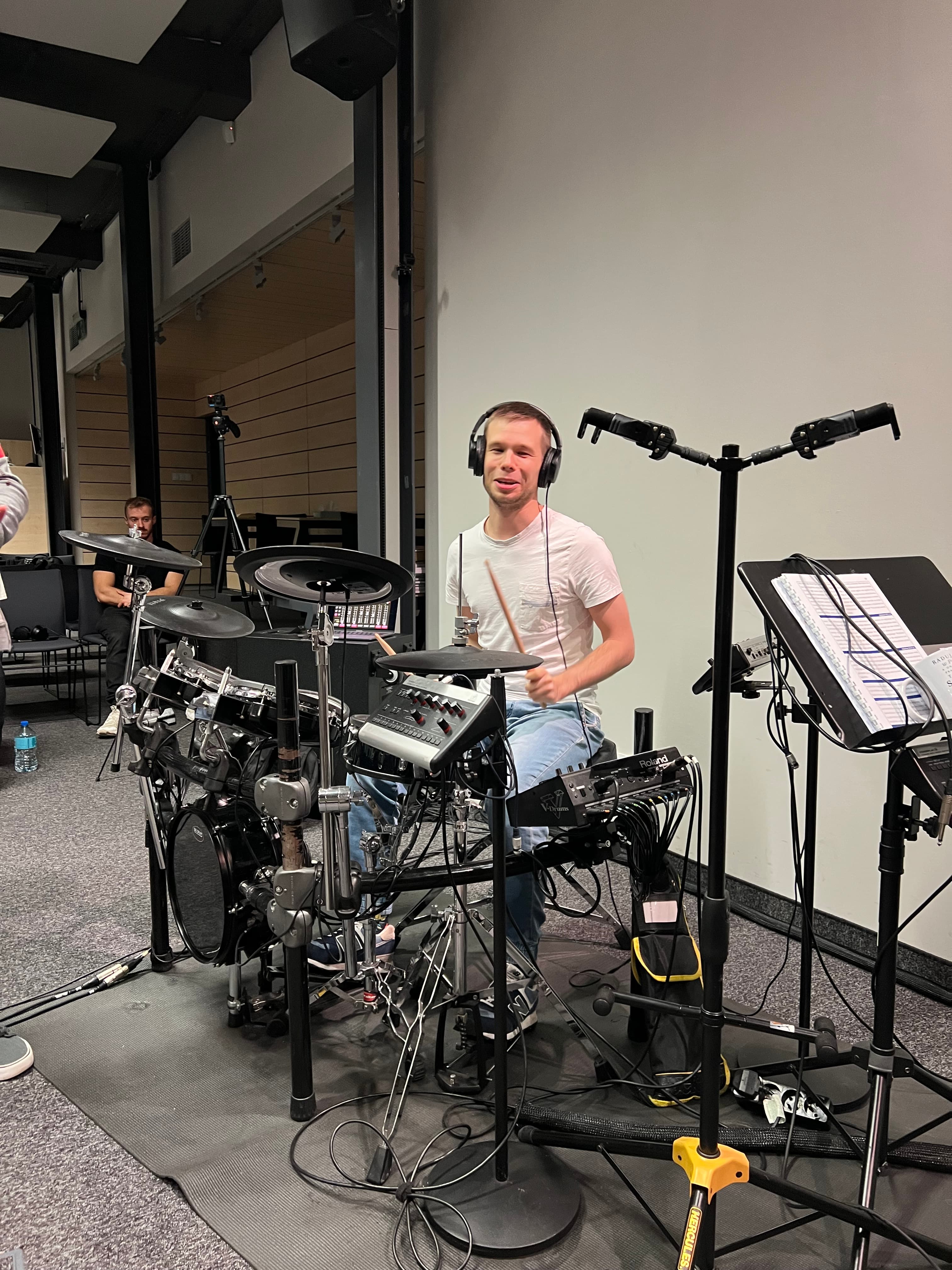 Obraz pokazuje młodego mężczyznę grającego na perkusji elektronicznej w studio nagraniowym. Ma na sobie słuchawki i białą koszulkę, a w tle widoczne są nuty, mikrofony i krzesła.