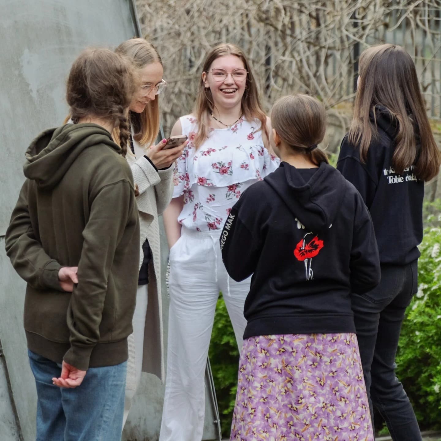 Obraz pokazuje pięciu młodych ludzi stojących na zewnątrz, rozmawiających i śmiejących się. Jedna osoba ma na sobie białą bluzkę z kwiatowym wzorem, a reszta ubrana jest w bluzy i kurtki. Tło obejmuje szarą ścianę i zielone krzewy.