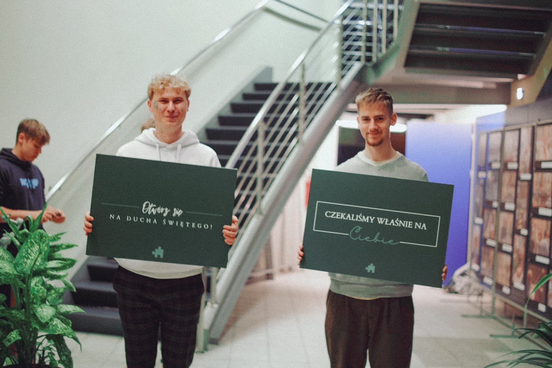 Obraz przedstawia dwóch młodych mężczyzn stojących w pomieszczeniu z klatką schodową, trzymających zielone tablice z napisami: po lewej stronie: "Otwórz się na Ducha Świętego", po prawej stronie: "Czekaliśmy właśnie na Ciebie". W tle widoczne są schody i rośliny.