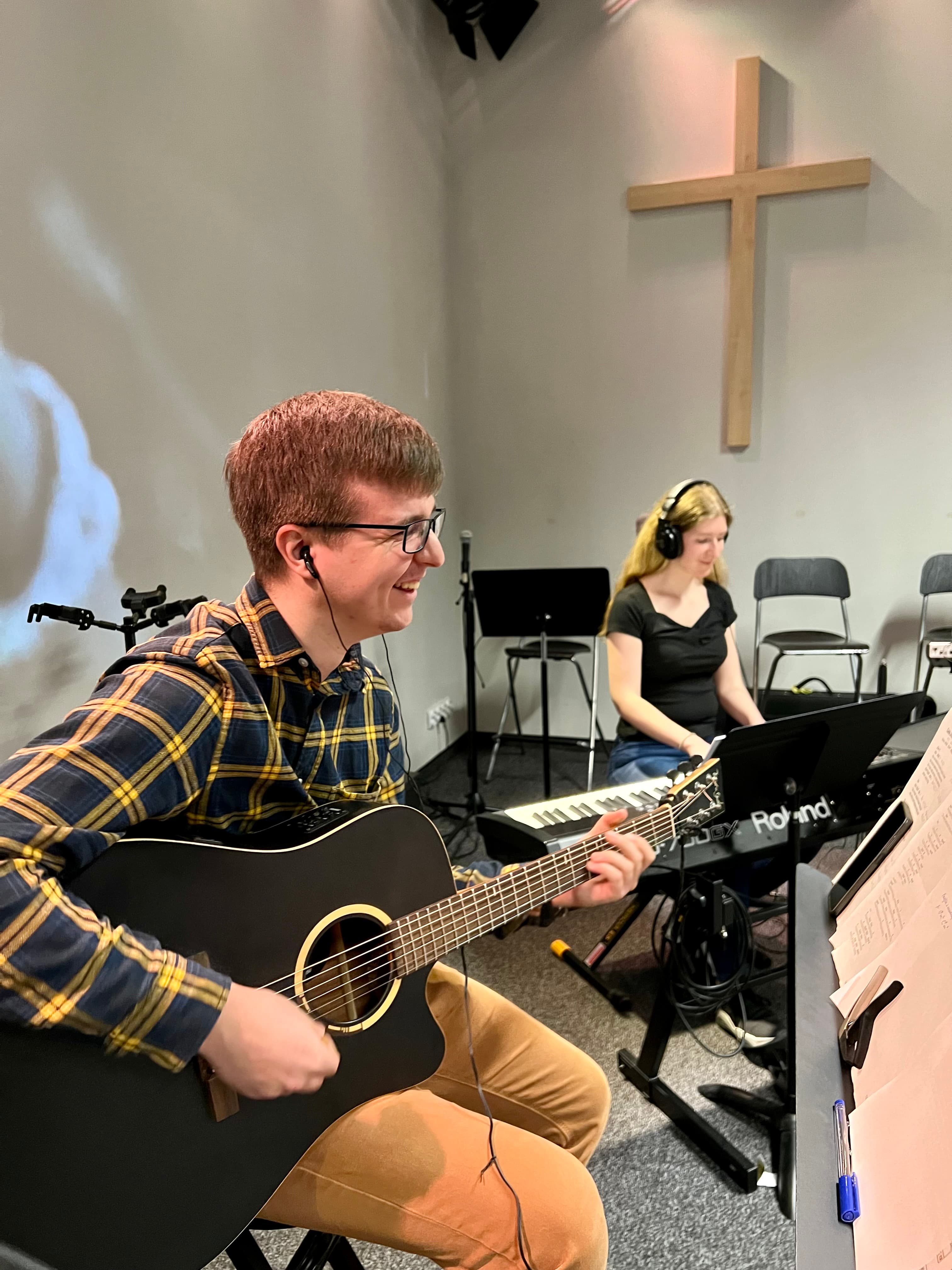 Obraz przedstawia młodego mężczyznę z gitarą i kobietę przy keyboardzie w studiu nagraniowym. Oboje mają słuchawki, a w tle widoczne są nuty, mikrofony i krzesła.