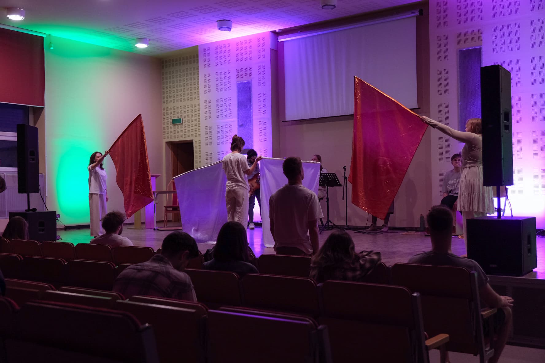 Grupa osób na scenie w sali z fioletowym i zielonym oświetleniem. Kilka osób macha czerwonymi i zielonymi flagami, a w tle widoczny jest ekran projekcyjny i głośnik. Publiczność siedzi na krzesłach i obserwuje wydarzenie