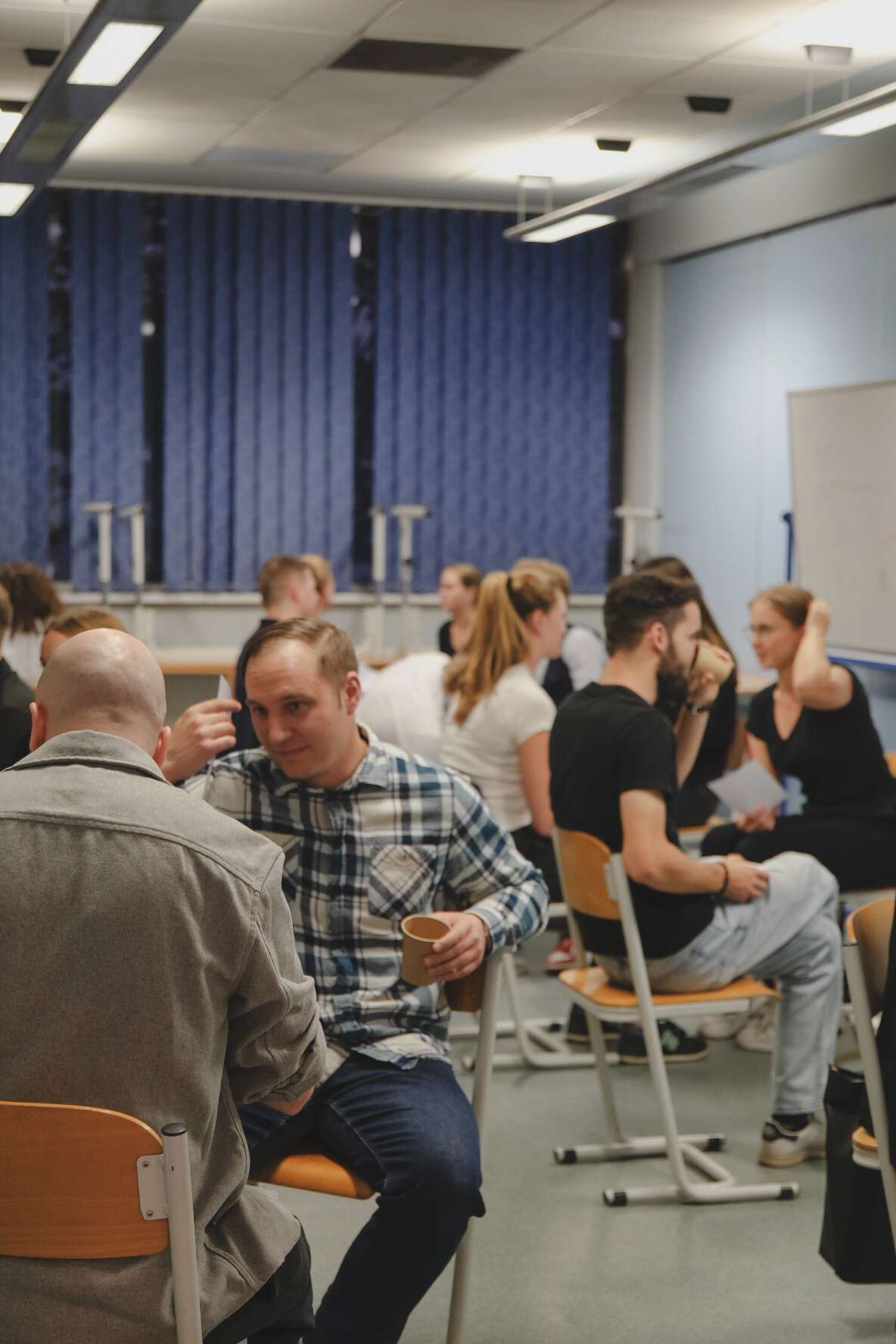 Obraz pokazuje grupę ludzi siedzących przy stole w sali z projektorem i ekranem w tle. Na stole znajdują się kubki, talerze z jedzeniem i butelka wody, a w rogu roślina dekoracyjna.