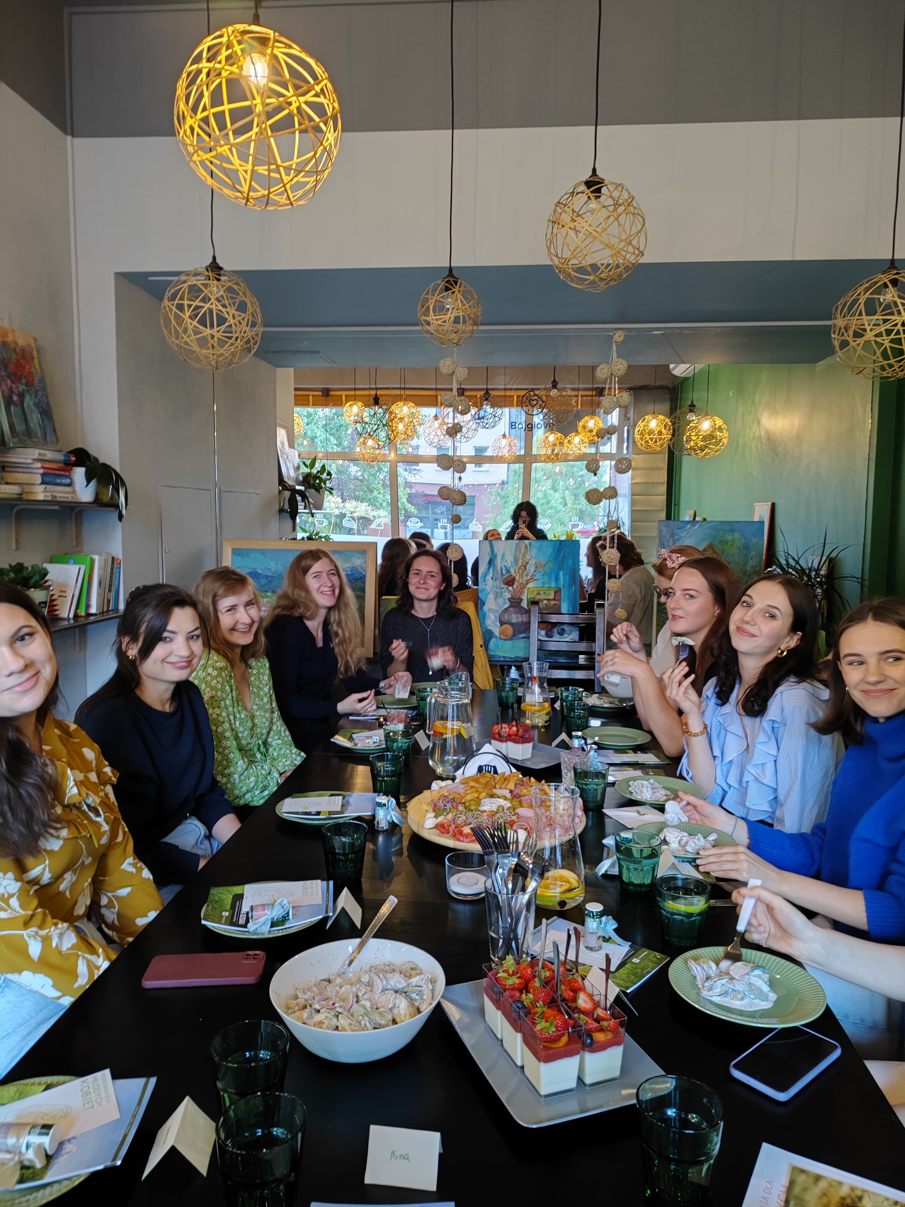 Grupa młodych kobiet siedzących przy stole w jasnym pomieszczeniu z dużymi oknami. Na stole znajdują się talerze z jedzeniem, szklanki, napoje i dekoracje, sugerujące wspólny posiłek lub spotkanie. W tle widoczne są obrazy na sztalugach i półki z książkami, co wskazuje na artystyczną lub kreatywną atmosferę.